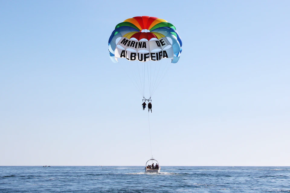 Two people parasailing over calm water, suspended from a colorful parachute labeled 'Marina de Albufeira,' with a boat below.