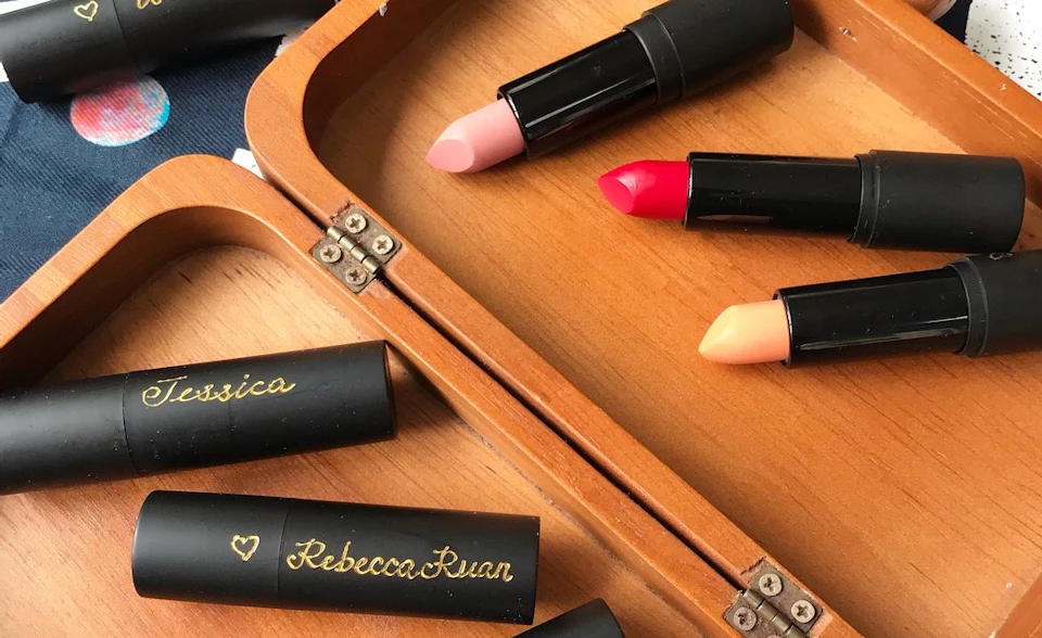Three lipsticks in pink, red, and peach shades are arranged on a wooden box, with two closed cases labeled "Rebecca Ruan."