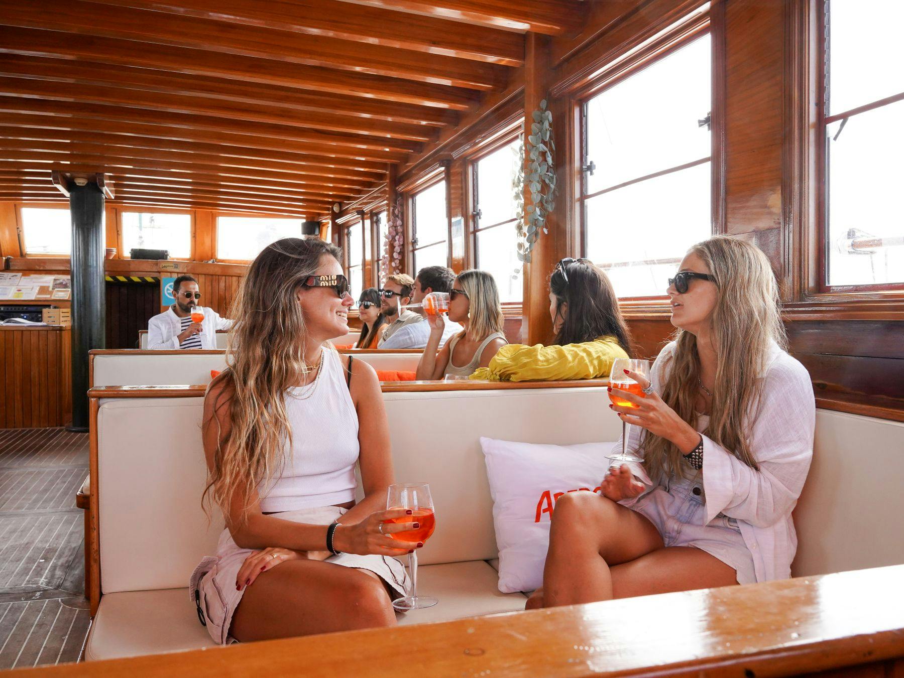 Deux femmes assises sur le banc rembourré d'un bateau, tenant des boissons et discutant. Un autre groupe de personnes est visible à l'arrière-plan.