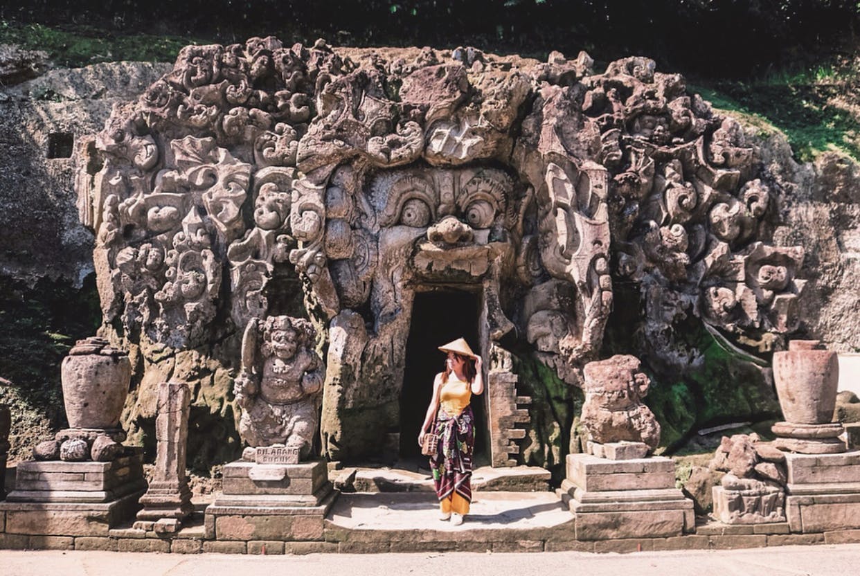 A person stands in front of a stone temple entrance, decorated with intricate carvings and statues, wearing traditional attire.