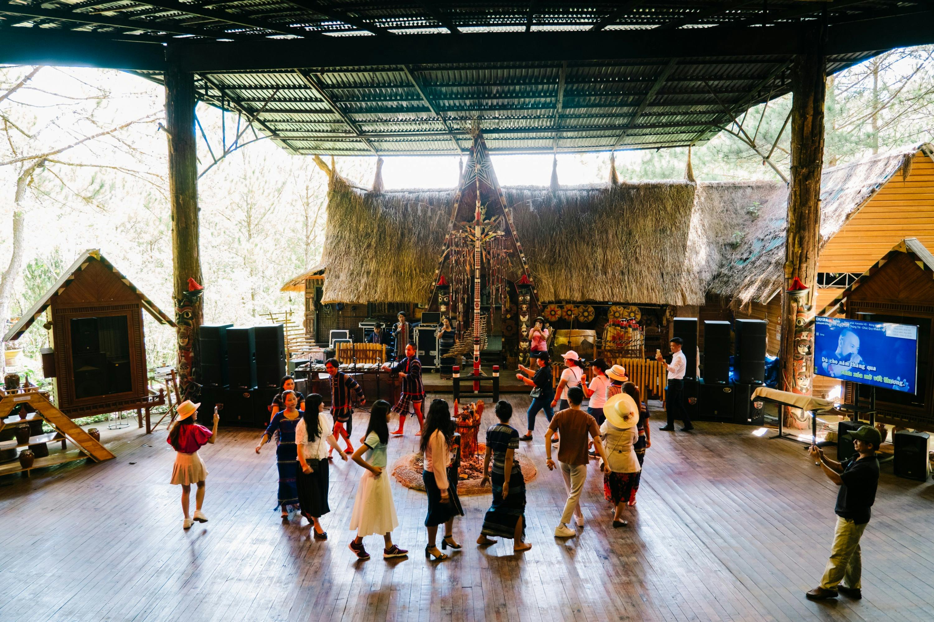 A group of people dancing in a rustic open-air venue with wooden floors and a thatched roof, decorated with cultural elements.