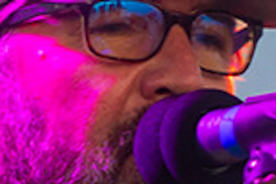 A musician wearing a floral shirt performs with an acoustic guitar and microphone on a waterfront stage under colorful lights.