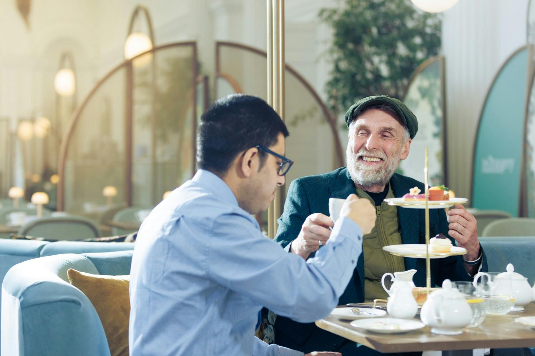 Two men sitting in a café, enjoying tea and pastries from a tiered tray. One man wears glasses, the other a green cap and smiles.