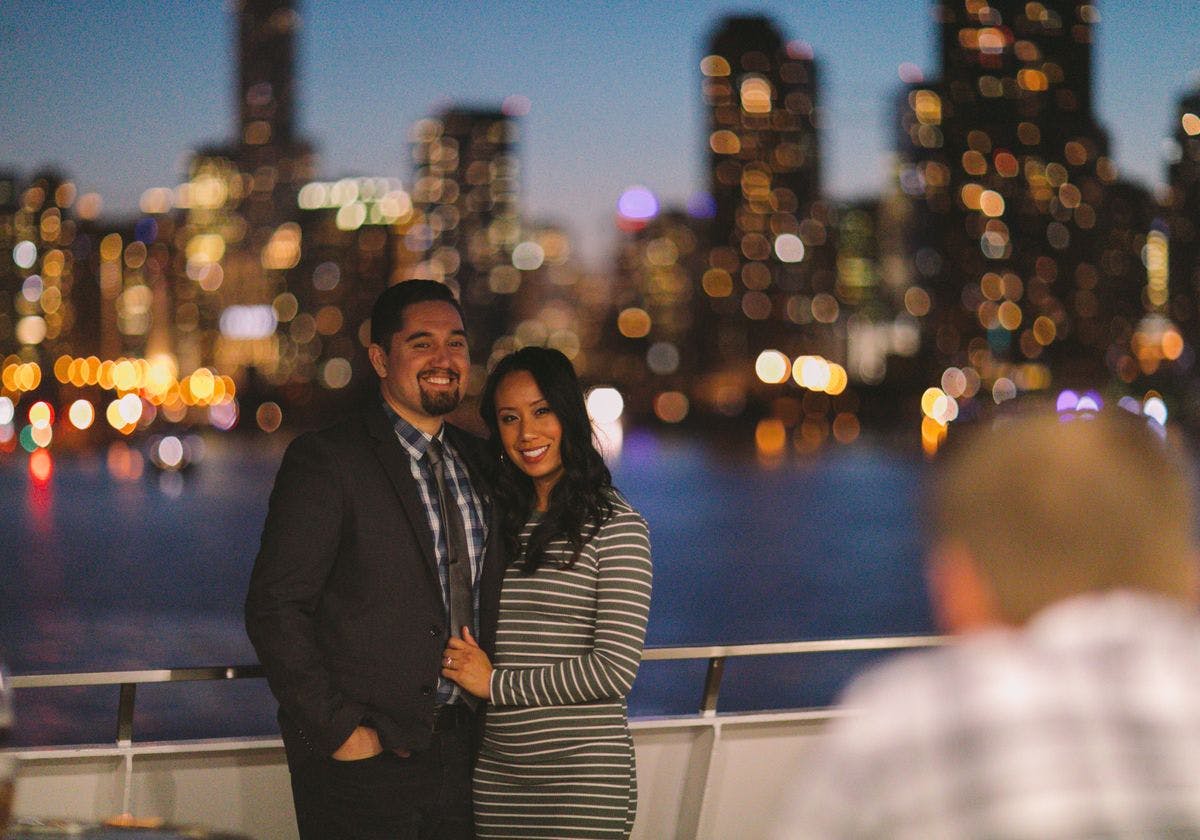 A couple standing on a boat at night with blurred city lights in the background. Both are smiling at the camera.