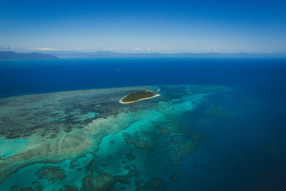 Aerial view of a small, lush island surrounded by turquoise waters and coral reefs under a clear blue sky.