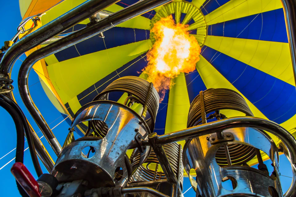 Close-up of a hot air balloon burner system igniting a flame, with the vibrant yellow and blue balloon canopy in the background.