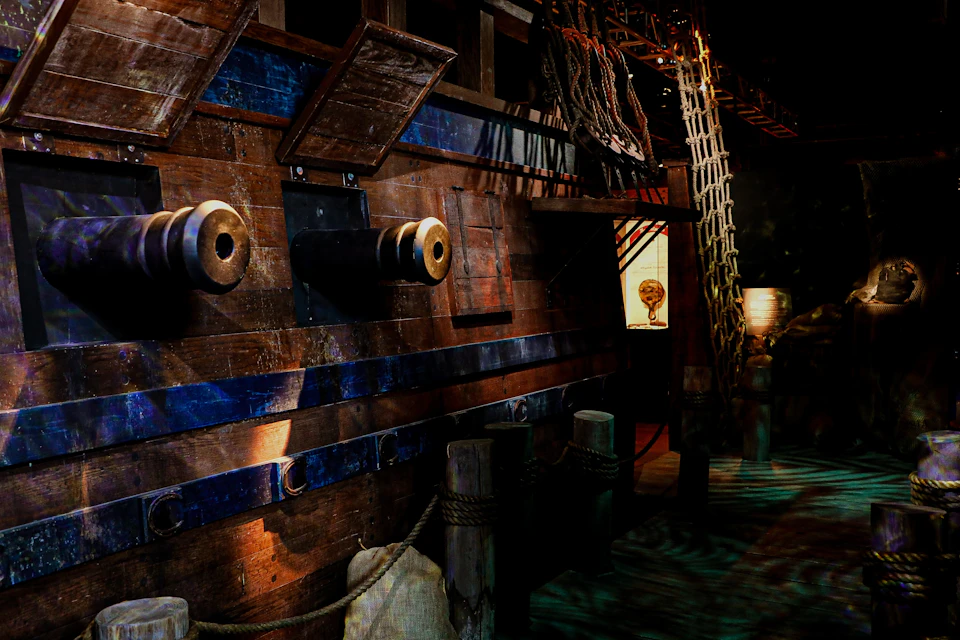 Wooden pirate ship with cannons, ropes, netting, and a lantern, docked at a pier with posts and ropes in dim lighting.