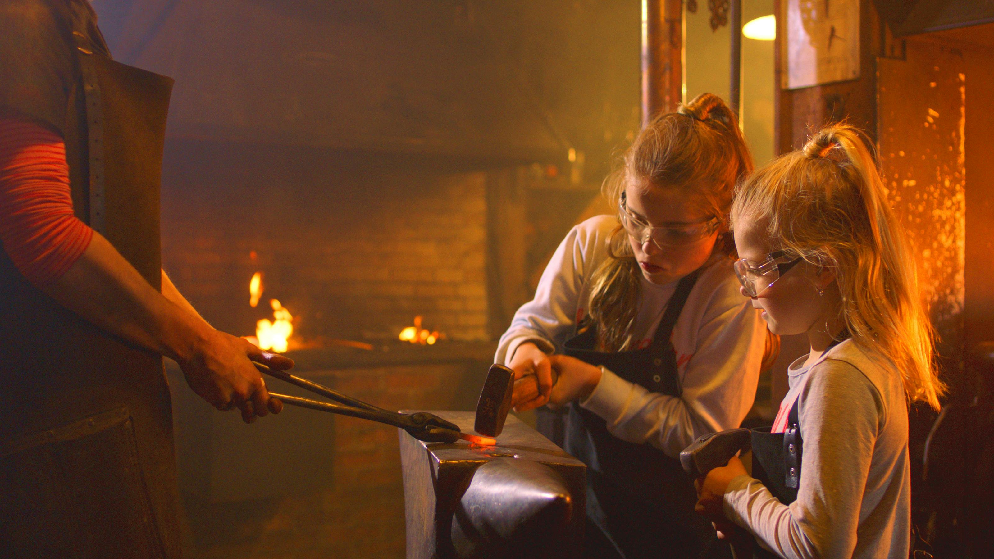 Dvě mladé dívky, které nosí ochranné brýle a zástěry, buší do kovu v tlumeně osvětlené dílně.