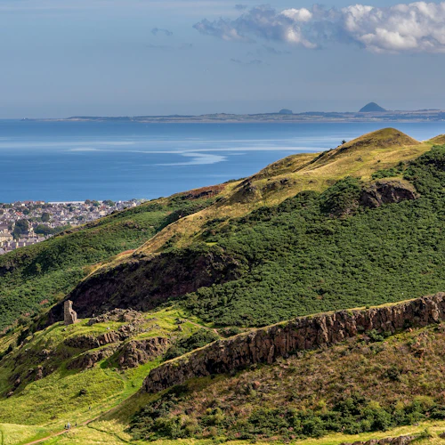 En naturskön utsikt över det gröna, kuperade landskapet som leder ner till en kuststad, med det blå havet och det avlägsna landet vid horisonten.