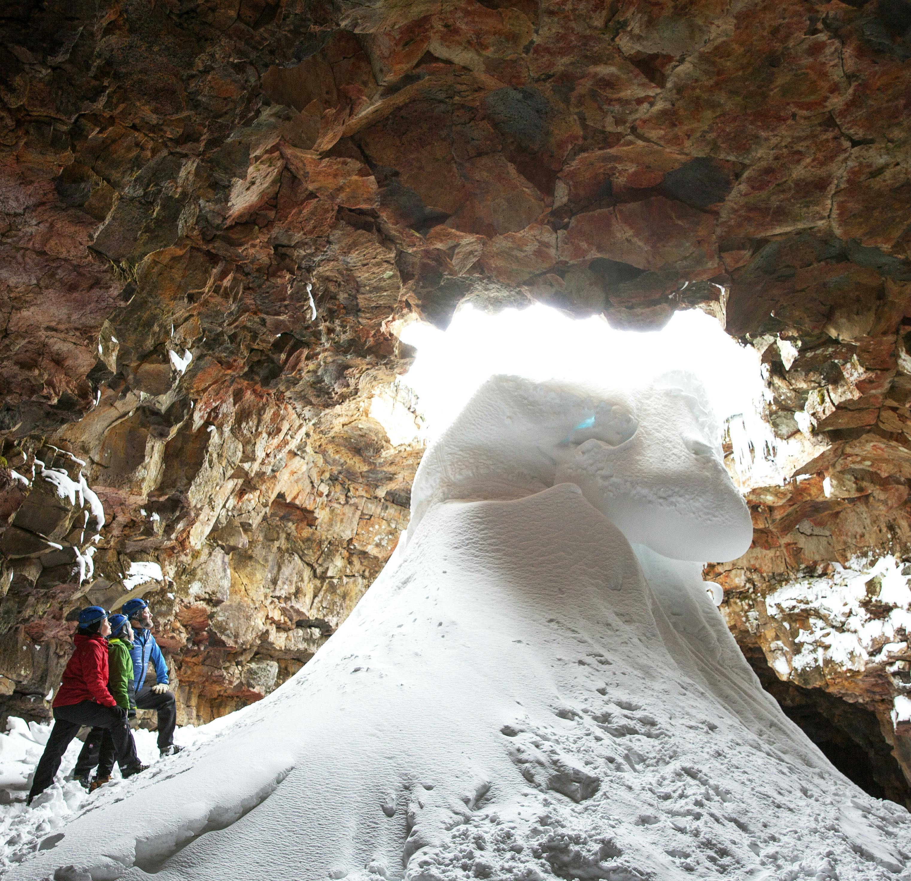 Three people in winter gear stand beside a large mound of snow inside a rocky, sunlit cave with a partially snow-covered entrance.