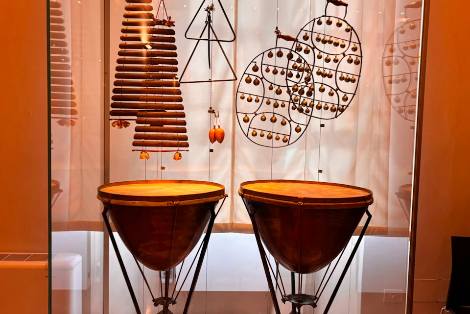 A display of percussion instruments, including two timpani and various hanging chimes and bells, in a well-lit setting.