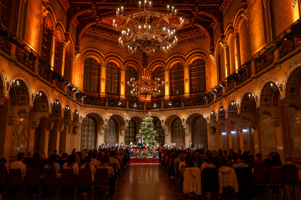 An ornate hall with chandeliers, a Christmas tree, and a seated audience watching an orchestra perform on stage.