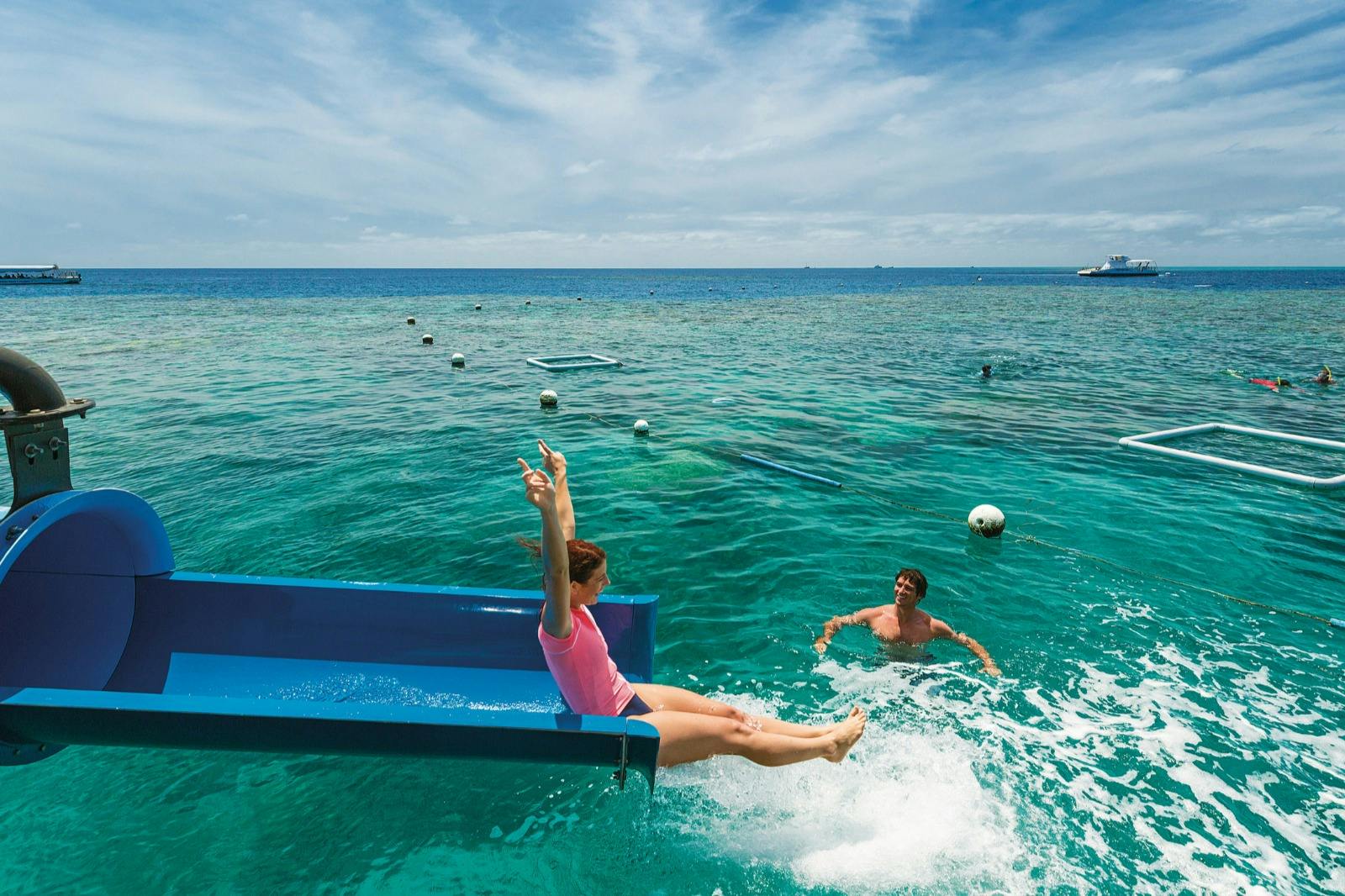 A person slides into the ocean from a blue slide, while another person nearby is in the water. The sky is partly cloudy.