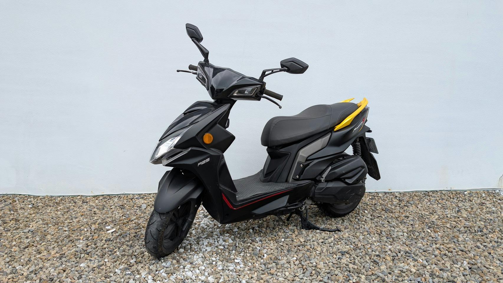 A black and yellow motor scooter parked on a gravel surface against a white wall.