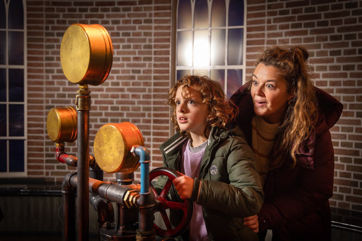 レンガの壁とアーチ型の窓がある部屋で、赤い車輪を備えた機械装置を操作している子供と大人。