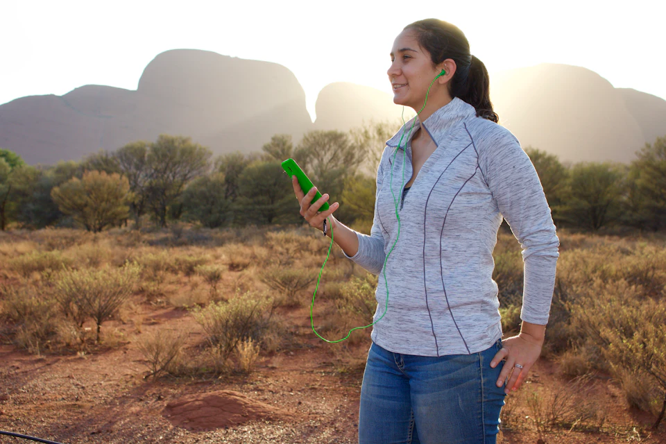 Person outdoors, wearing earbuds, holding a smartphone, smiling, with a desert landscape and distant rock formations in the background.