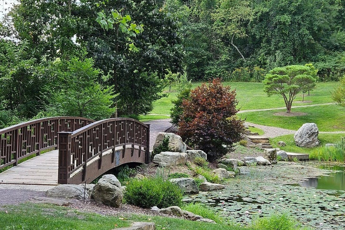 Un parc pittoresque avec un petit pont en bois au-dessus d'un étang entouré de verdure, d'arbres, de rochers et d'un sentier de promenade.