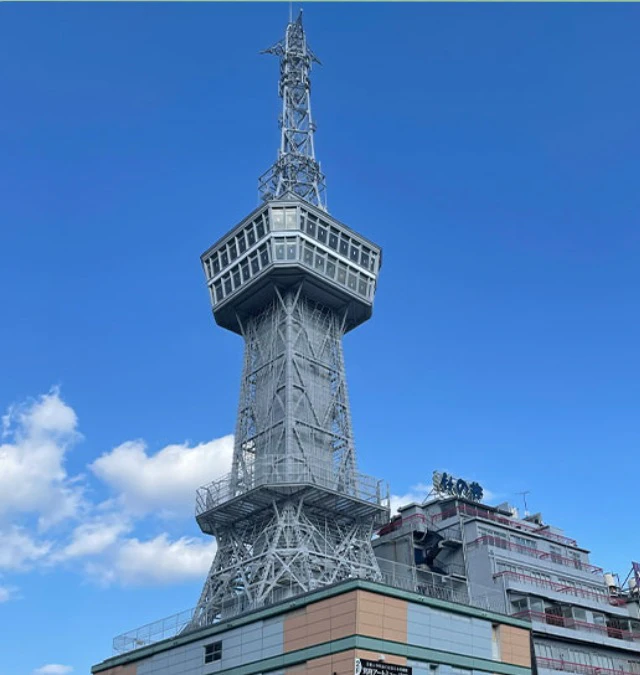 A tall metallic tower with an observation deck and antenna rises against a blue sky, with nearby buildings visible.
