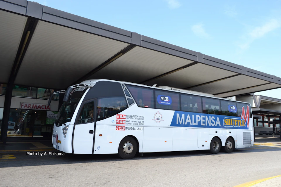 A white shuttle bus labeled "MALPENSA" is parked under a large roof structure with clear skies in the background.