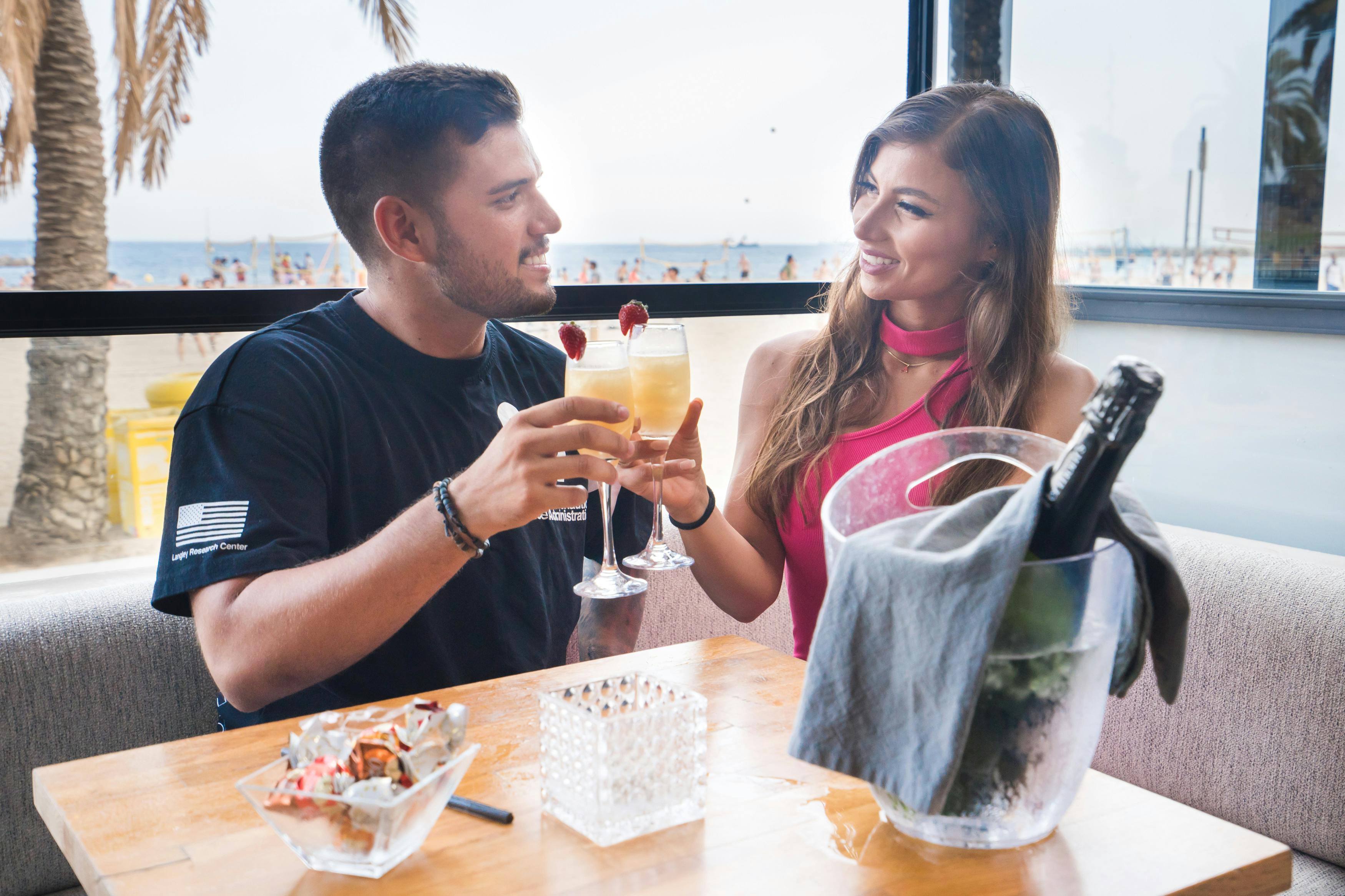 Ein Mann und eine Frau stoßen an einem Strandtisch mit einem Cocktail an, während im Hintergrund Palmen und das Meer zu sehen sind.
