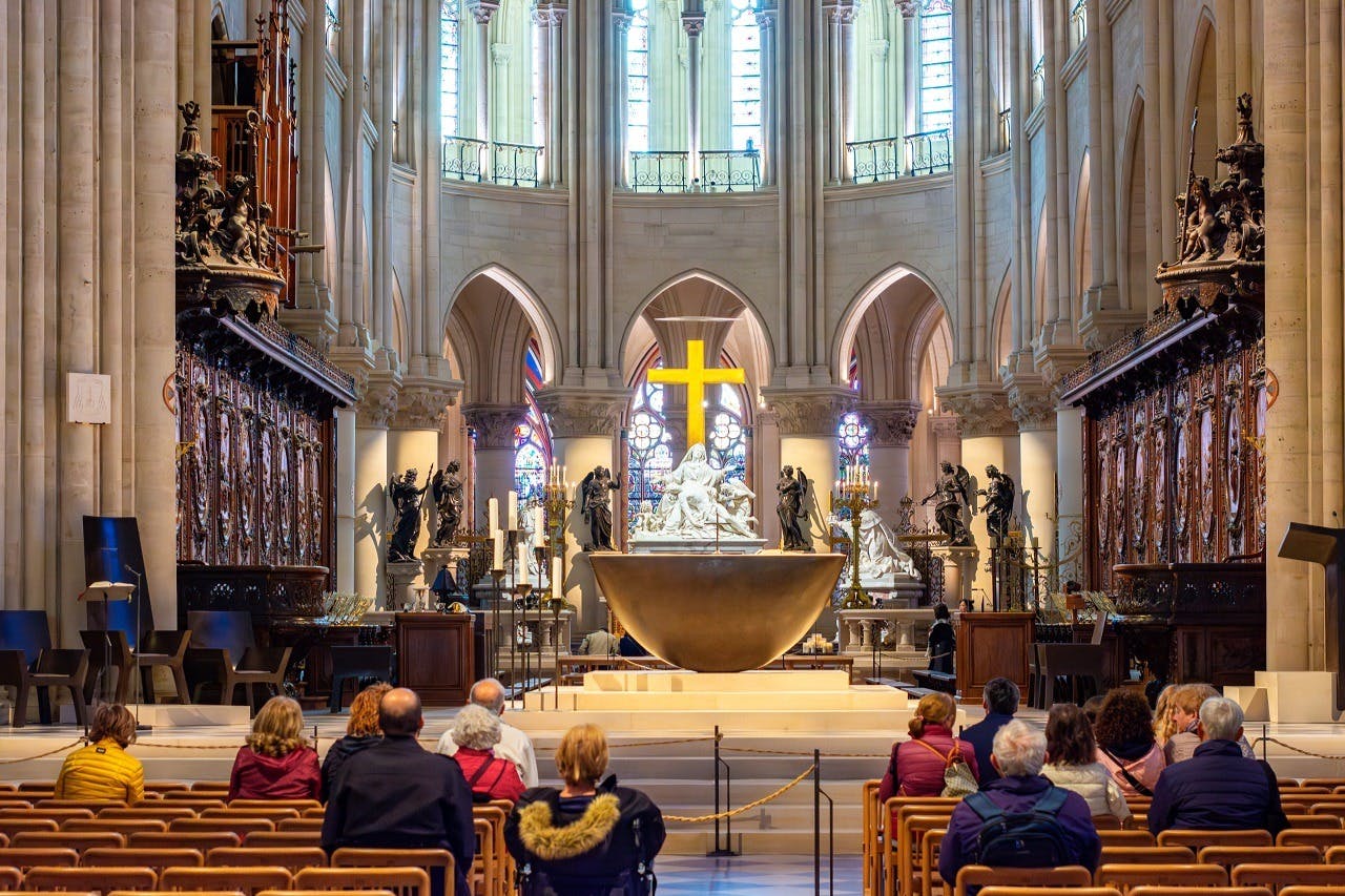 People seated in a church, facing a large altar adorned with a cross and statues. Stained glass windows are visible in the background.