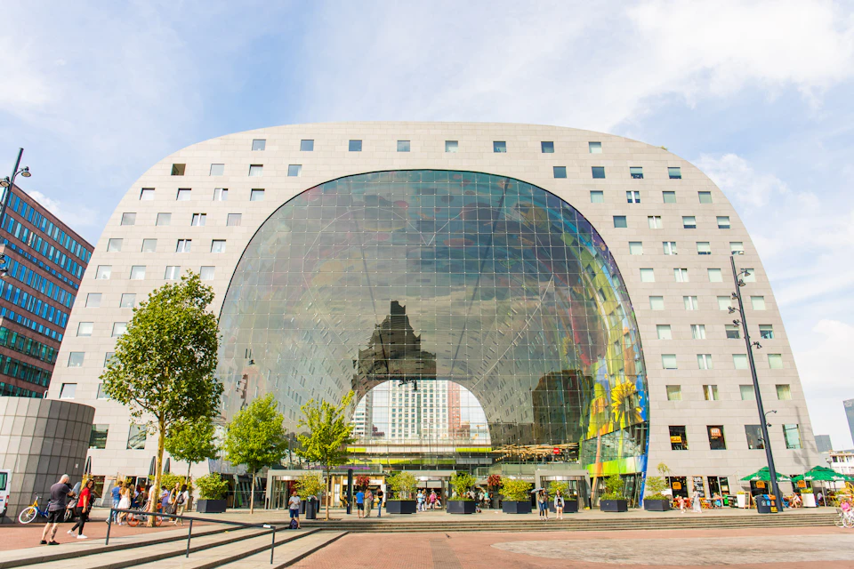 A modern, arched building with a reflective glass facade and many small windows. People are walking in front of it. Trees and potted plants line the entrance.