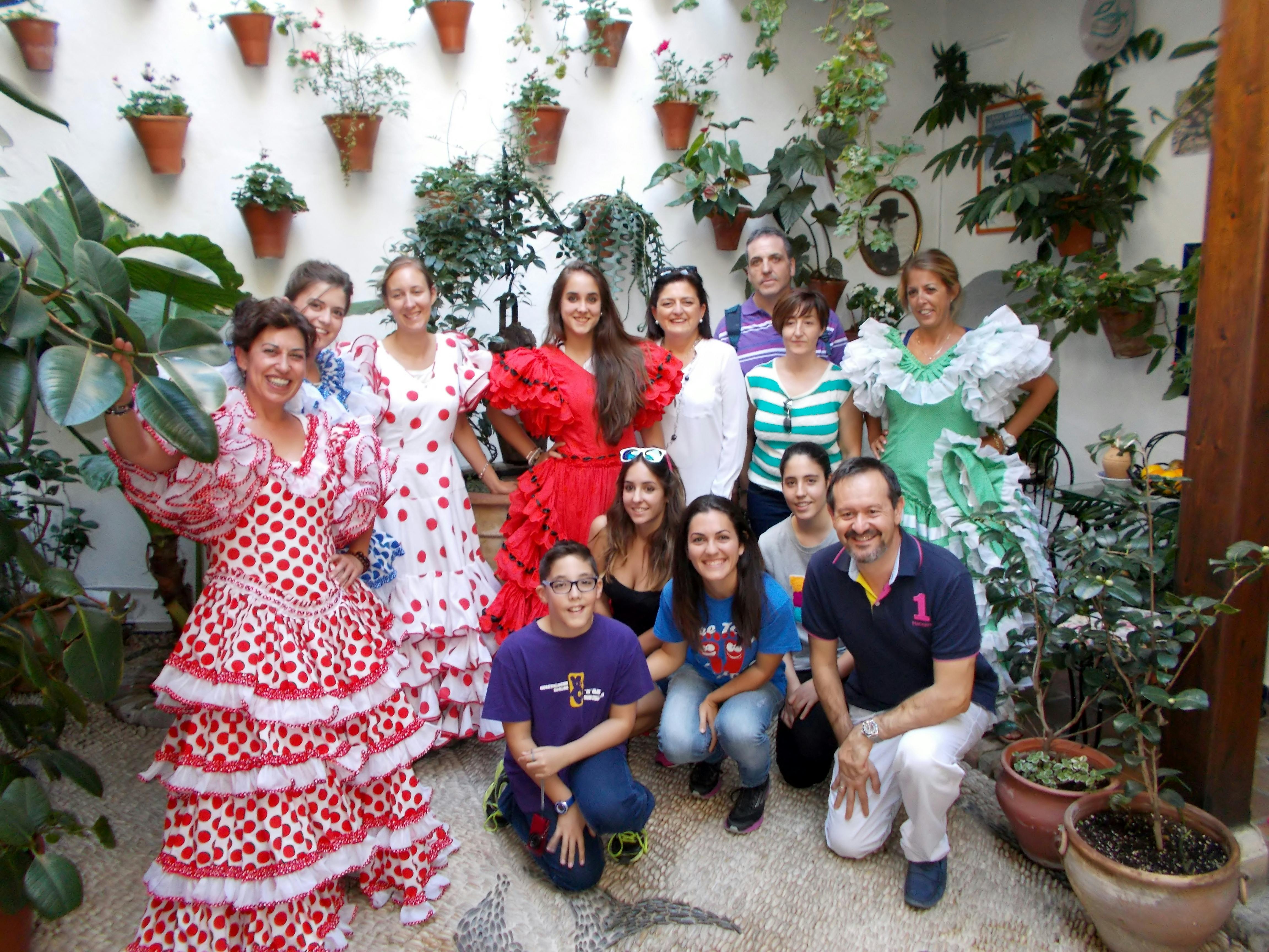 Group of people posing in colorful traditional attire with potted plants on a white wall in the background.