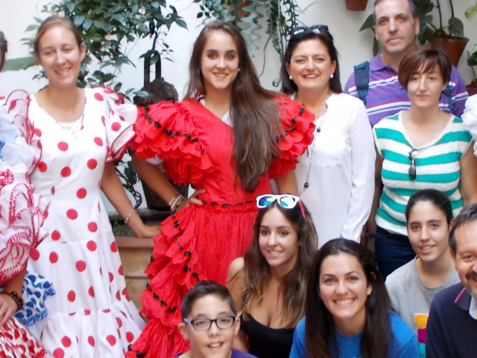 Group of people posing in colorful traditional attire with potted plants on a white wall in the background.