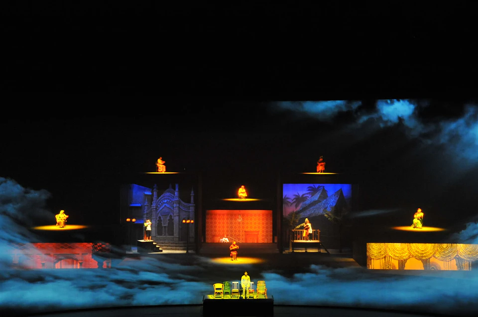 A stage performance featuring colorful, multi-level sets and actors in various costumes, illuminated by dramatic lighting.