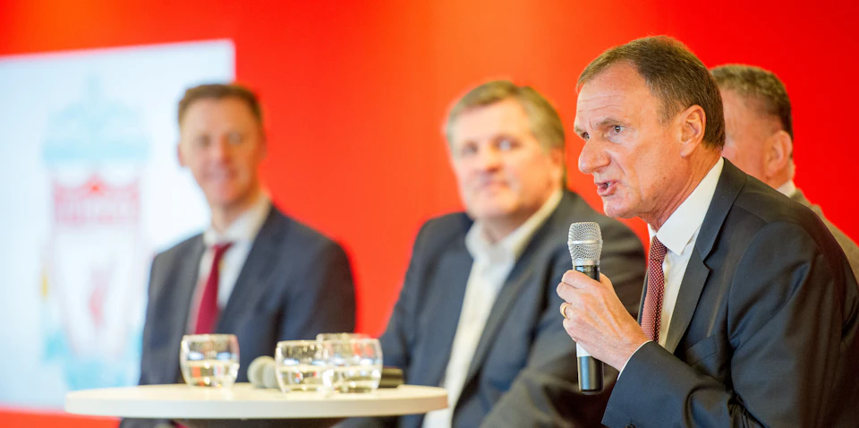 Man in a suit speaking into a microphone, with a blurred background featuring another man and a red wall.