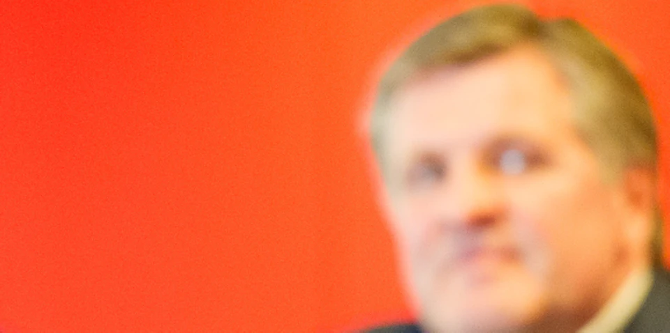 Man in a suit speaking into a microphone, with a blurred background featuring another man and a red wall.