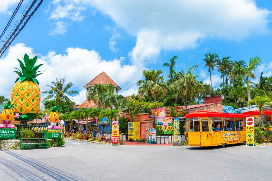 A colorful outdoor market scene with palm trees, a yellow tram, and a building with a red roof under a blue sky.