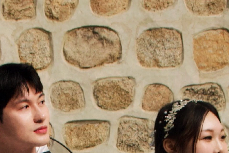 A couple wearing traditional clothing sits on a bench in front of a stone wall with plants.