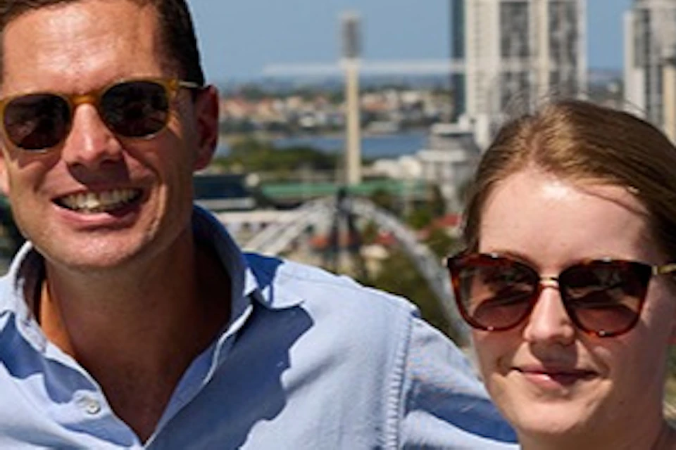 Two people posing for a photo on a scenic overlook with a cityscape and a river in the background under a clear sky.