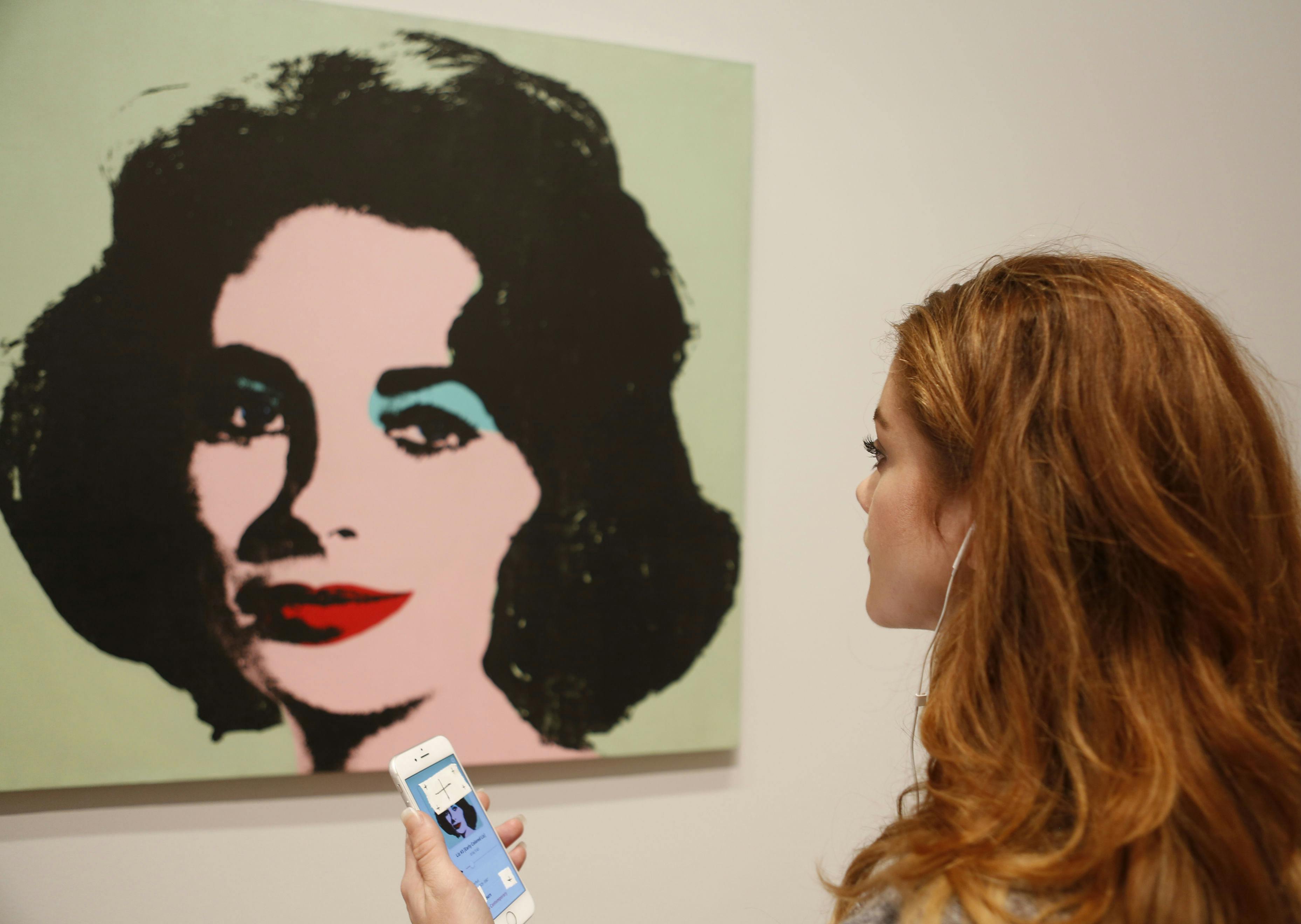 Una persona tiene in mano uno smartphone e guarda il dipinto di una donna con labbra rosse e ombretto blu su uno sfondo verde chiaro.