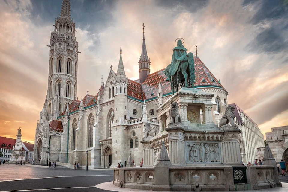 Gothic church with ornate rooftops, tall spires, and a statue in front at sunset. Few people walking nearby.