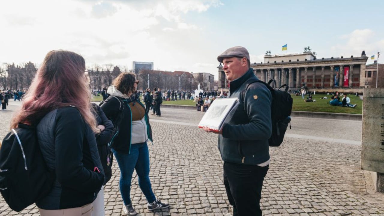 A man holds artwork and stands on a cobblestone square. People are in the background near a historic building with a Ukrainian flag.