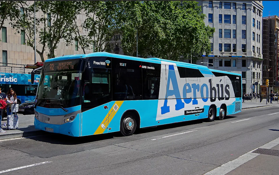 Blue and white bus labeled "Aerobús" parked on a street, offering free WiFi, with buildings and trees in the background.