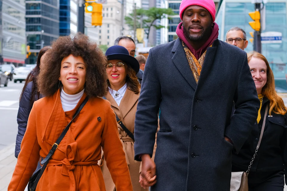 A group of people walking on a city sidewalk on a bright day; the central individuals are wearing colorful coats.