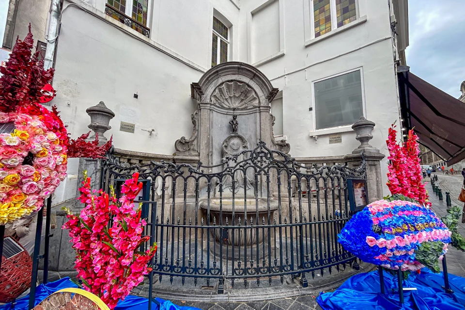 A decorative stone fountain with a small statue, surrounded by a black wrought iron fence and colorful floral arrangements.