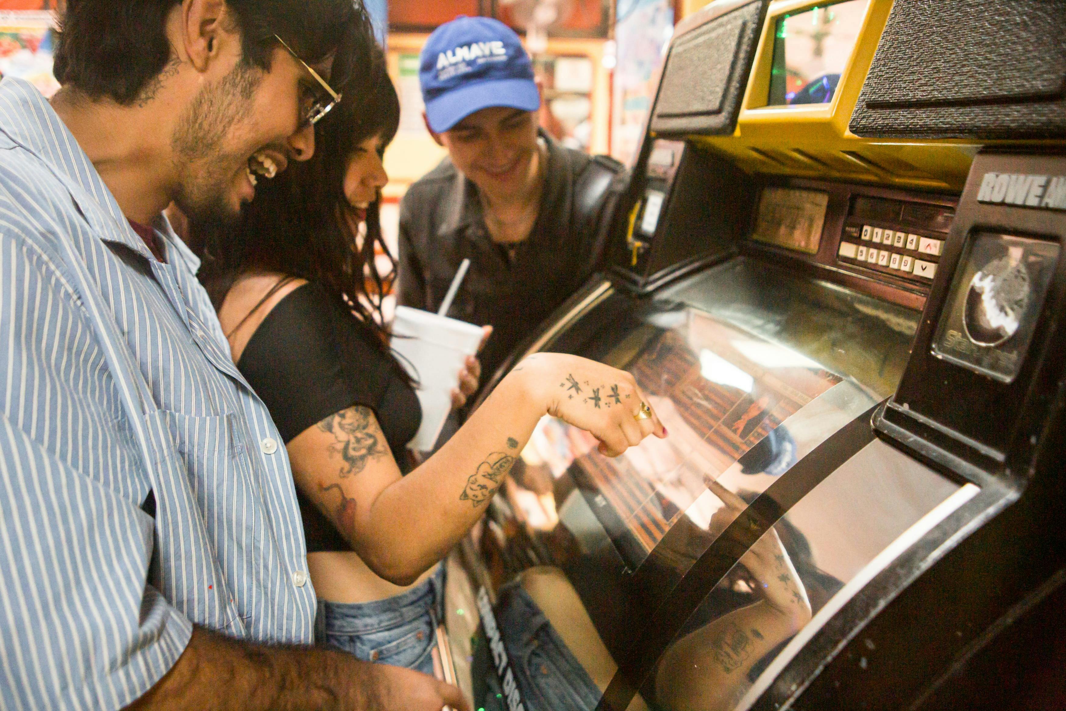 Drei Personen interagieren mit einer Jukebox in einer lebhaften Umgebung, lächeln und beschäftigen sich mit dem Gerät.
