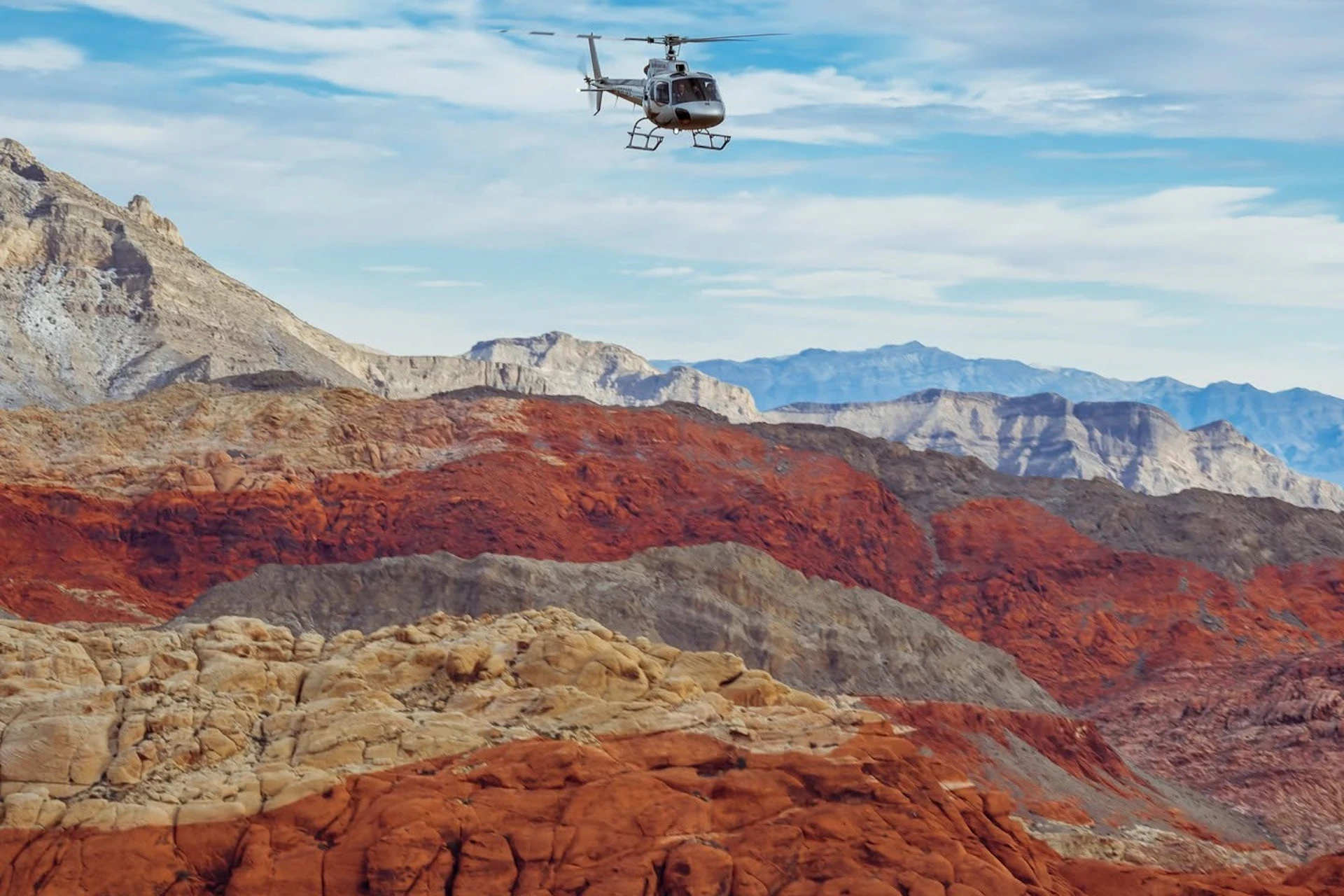 A helicopter flies over a landscape of colorful layered rock formations under a partly cloudy sky.