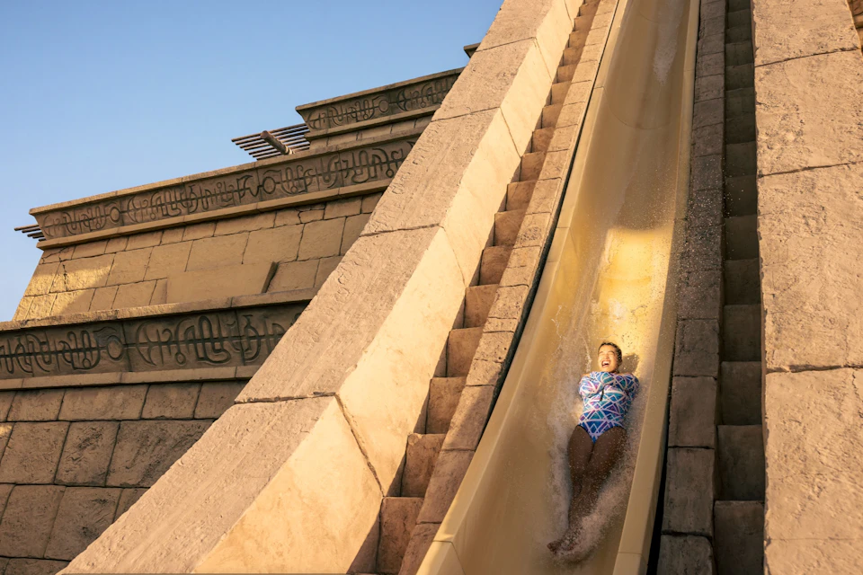 A person sliding down a steep water slide at a water park, surrounded by stone-like structures, under a clear sky.