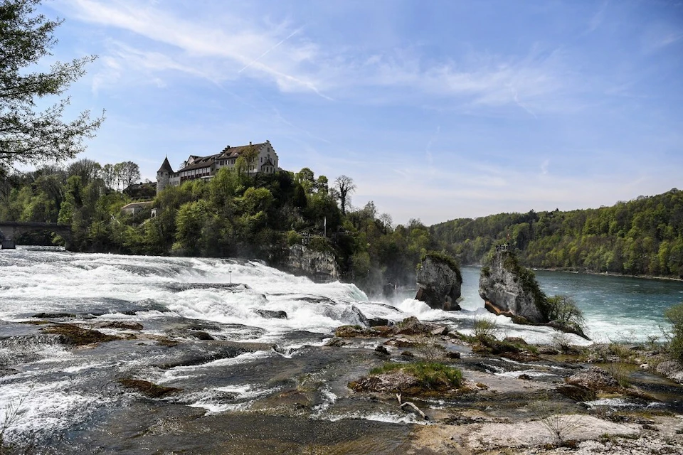 A scenic view of a large waterfall with a historic building on a hill surrounded by greenery under a clear blue sky.