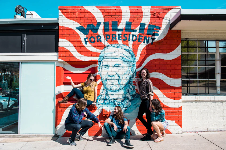 A group of people poses near a mural featuring a portrait and the text "Willie for President" on a red and white wall.