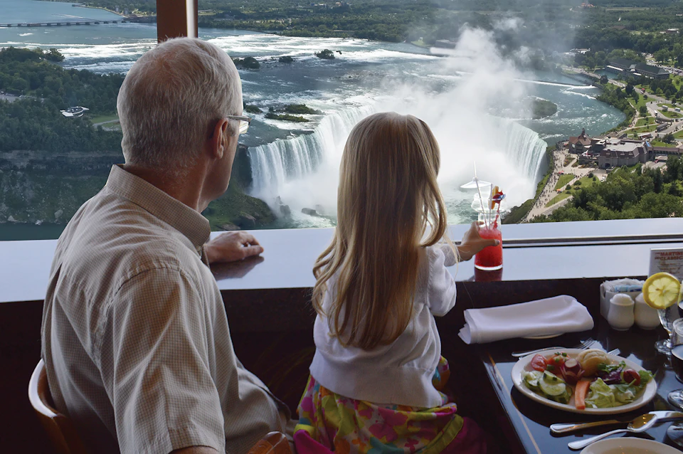 An older man and a young girl sit at a table overlooking a large waterfall, with the girl pointing at the view while holding a drink.