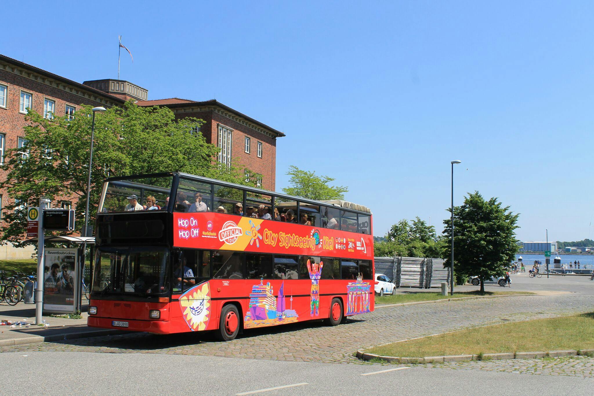 A red double-decker sightseeing bus filled with passengers, driving near a brick building and waterfront under a clear blue sky.