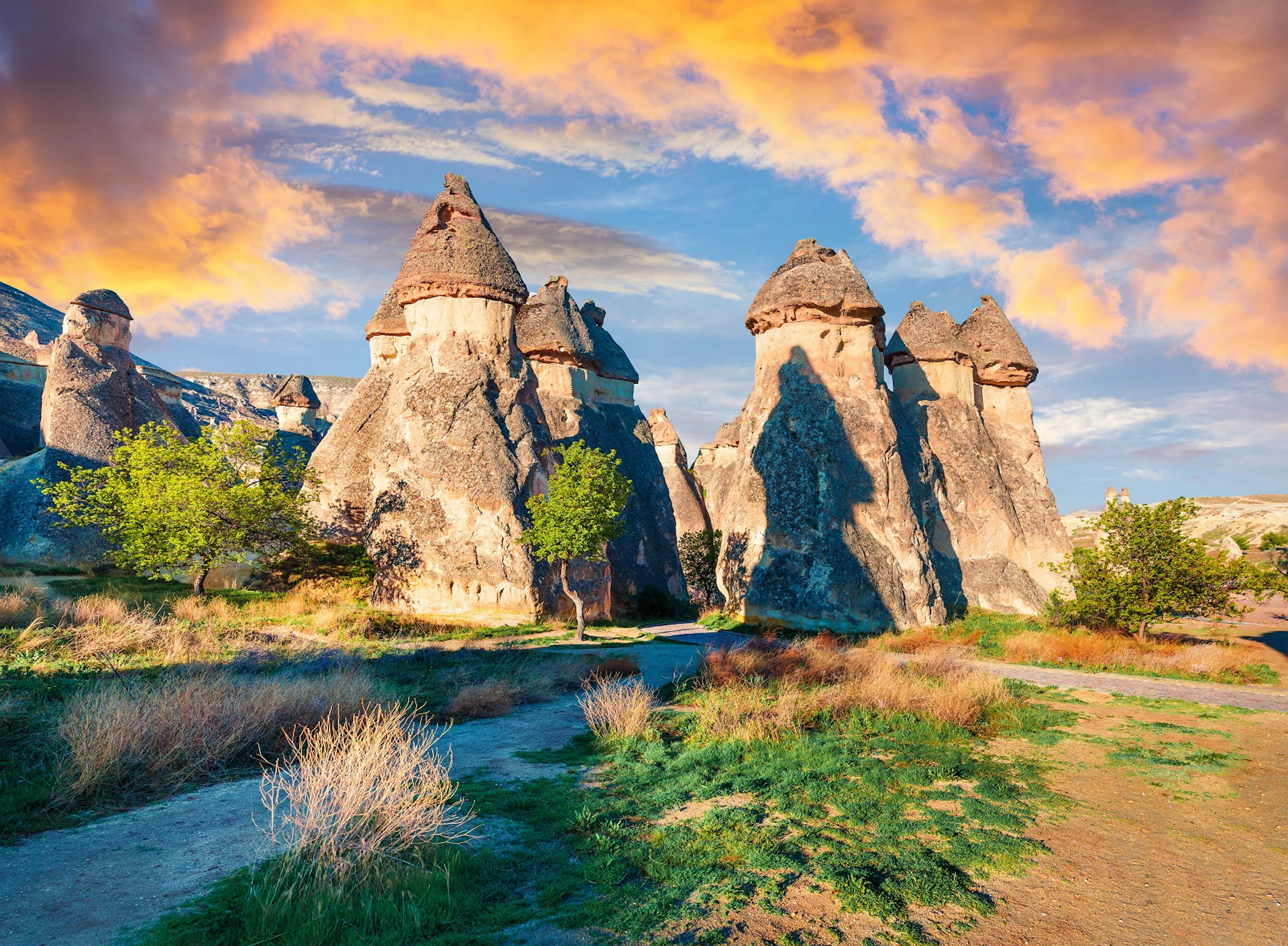 Unique rock formations with cone-shaped tops under a vibrant sunset sky, surrounded by greenery and a dirt path.