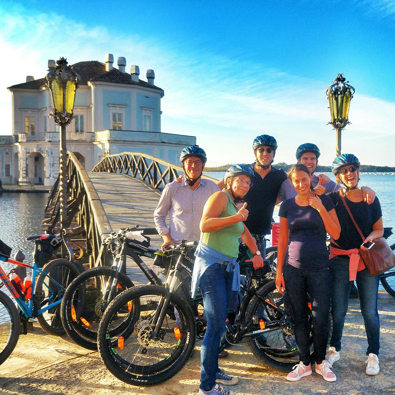 Six people in cycling gear pose with their bikes near a scenic bridge and a historic building by the water.