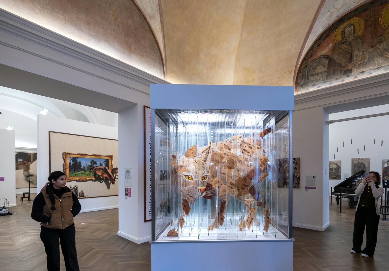 En kvinde står i et museumsgalleri med kunstudstillinger, herunder en geometrisk dyreskulptur i en glaskasse og malerier på vægge.